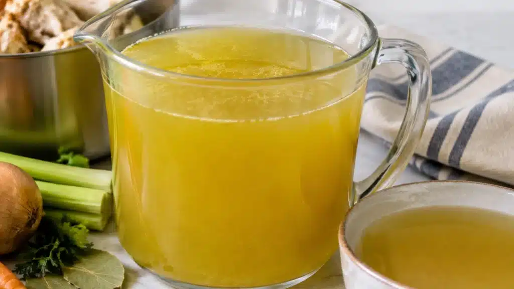 Homemade golden chicken stock in a glass jug, served beside a bowl with fresh celery, onion, herbs, and cooked chicken pieces.