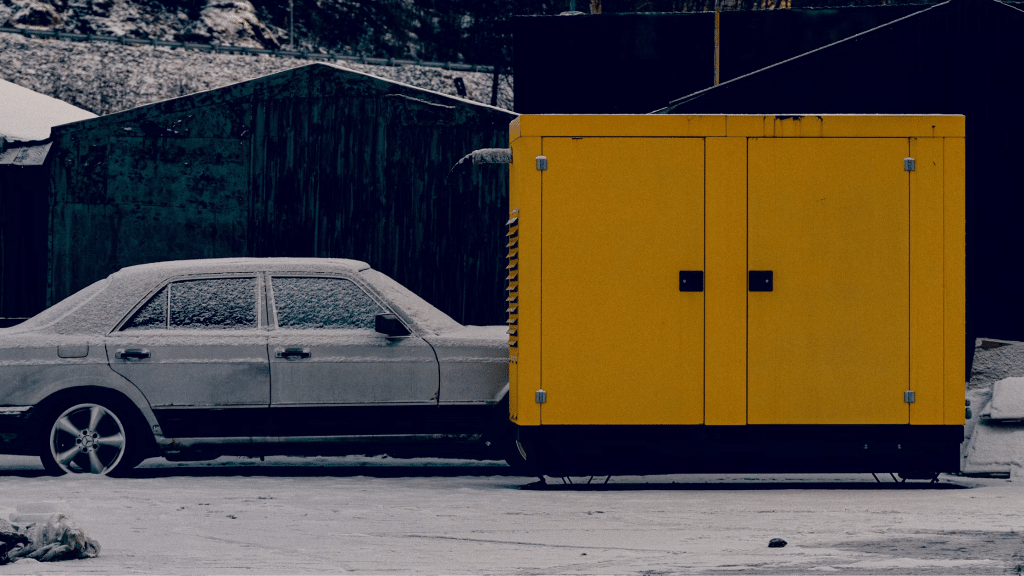 Snow-covered car parked beside large yellow generator outdoors in winter setting