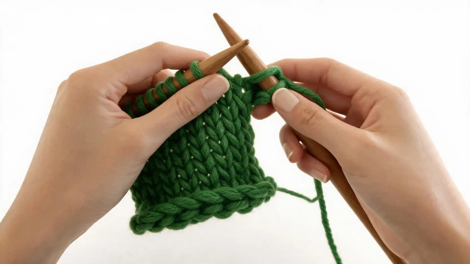 Hands knitting the first two stitches with green yarn and wooden needles to begin casting off.