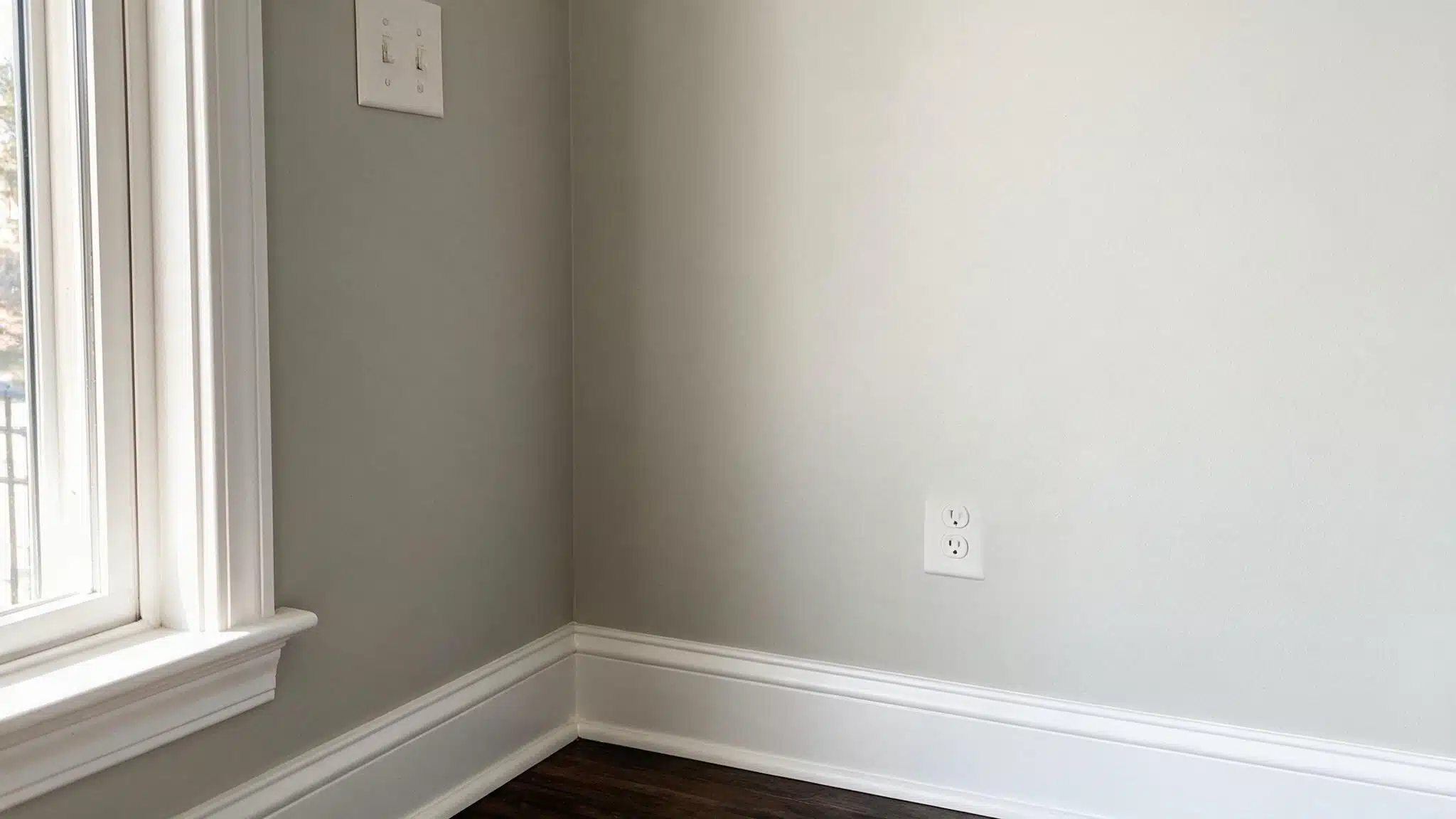 Empty room corner with gray walls, white trim and baseboards, a window on the left, two wall outlets, and dark wood flooring.