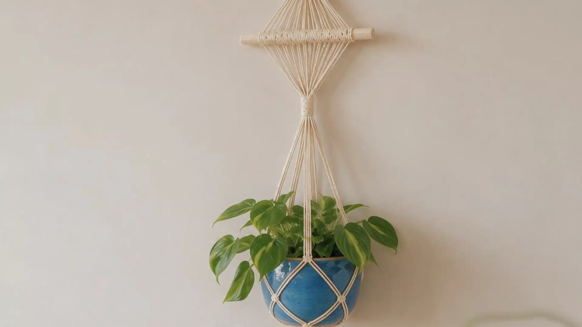 Cream tassel plant hanger holding a blue ceramic pot with a leafy green plant against a plain beige wall.
