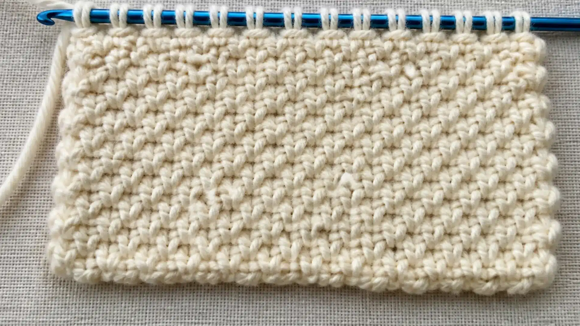 Pebbled reverse stitch texture stands out across a cream swatch with a blue Tunisian hook along the upper edge.