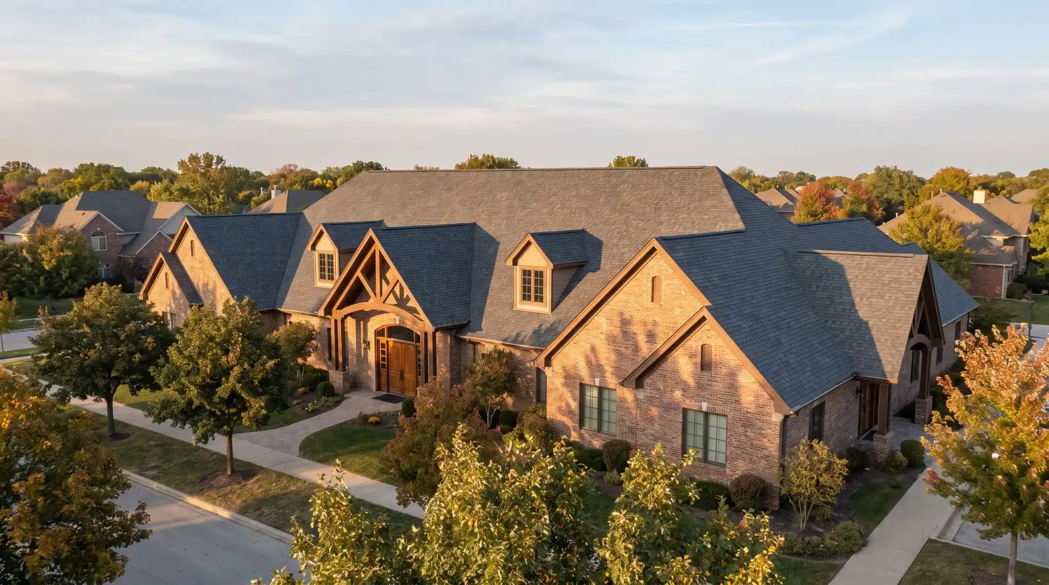 Large brick house with dark roof surrounded by trees in suburban neighborhood during sunset